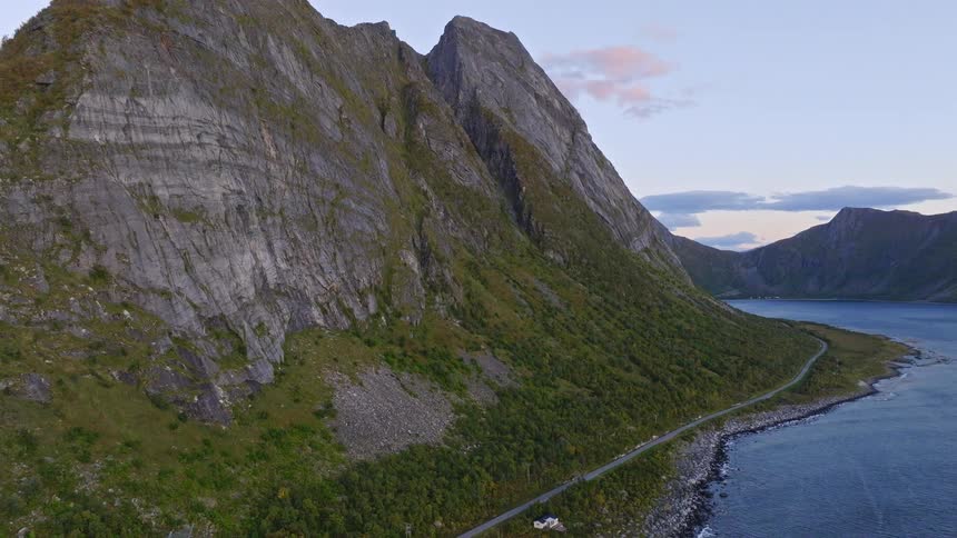 Stunning aerial pullback shot of a scenic road running along a fjord at the base of towering mountains