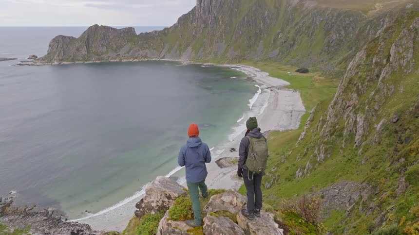 Aerial view reveals two hikers standing on a dramatic cliff, looking out over a pristine, secluded beach and the ocean in Norway