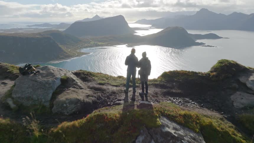 Aerial drone shot pulls back to reveal a couple standing on a mountain summit, enjoying the breathtaking view of a shimmering bay surrounded by majestic mountains