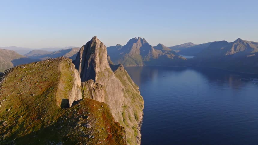 Breathtaking aerial footage pulls back over the jagged ridge of a majestic mountain, revealing a stunning Norwegian fjord landscape under a clear blue sky