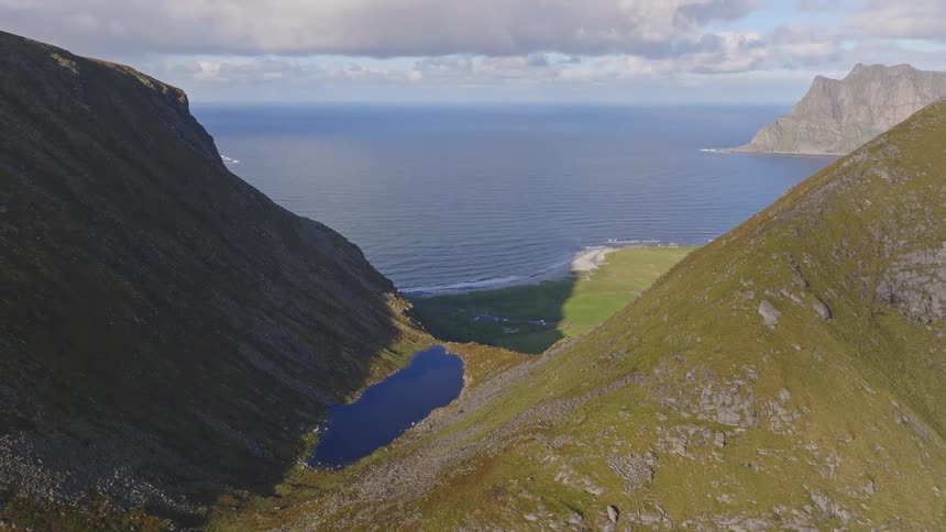 A majestic aerial pullback reveals a stunning mountain valley with a tranquil lake leading out to the open sea under a partly cloudy sky