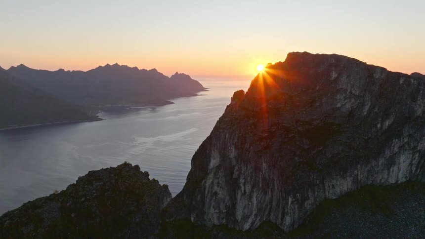 Epic aerial footage reveals the sun rising from behind a dramatic mountain, casting its warm glow over a scenic fjord