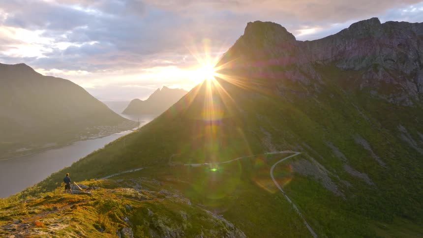 A lone hiker stands atop a mountain ridge, watching the sun crest over a majestic peak with an epic view of the surrounding fjords