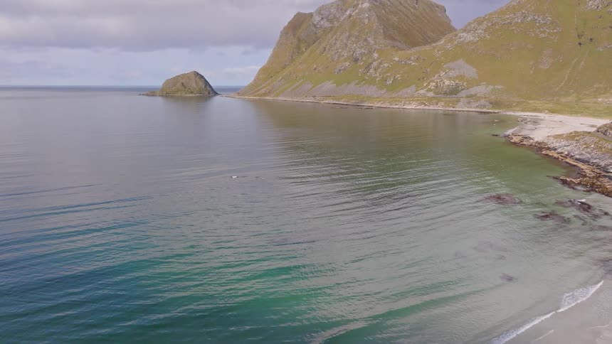 An aerial view of a serene beach with calm, clear water lapping at the shore, framed by majestic green mountains