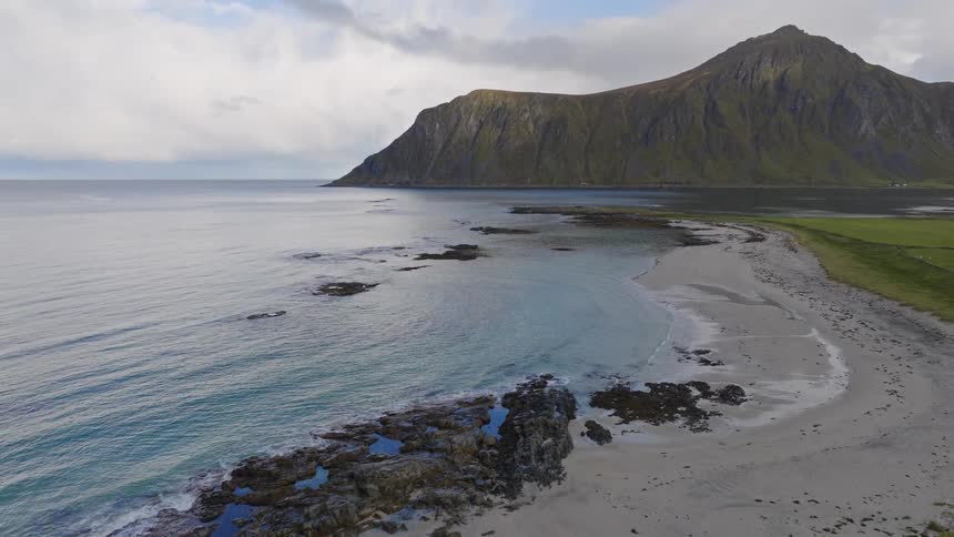 An aerial view slowly pulls back to reveal a majestic and remote beach with turquoise waters and a towering green mountain