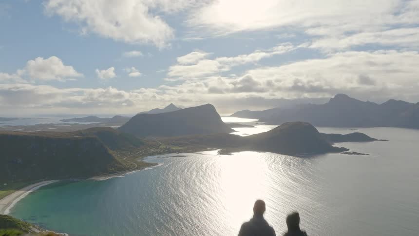 An aerial shot flies over two silhouetted hikers on a mountain peak to reveal a stunning vista of fjords and beaches below