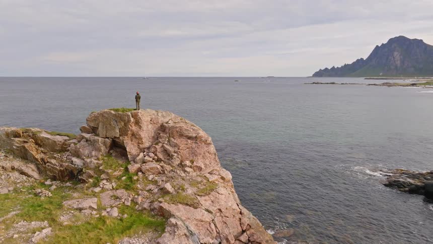 Aerial view reveals a lone traveler standing on a rocky precipice, gazing out at a majestic coastal landscape with distant mountains