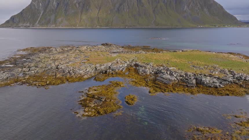 An aerial view pulls back from a rocky islet surrounded by clear water to reveal a massive, majestic mountain in the Lofoten Islands, Norway