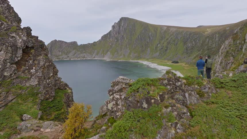 An aerial view pushes past a couple hiking on a cliff to reveal a stunning secluded beach nestled in a dramatic mountain landscape