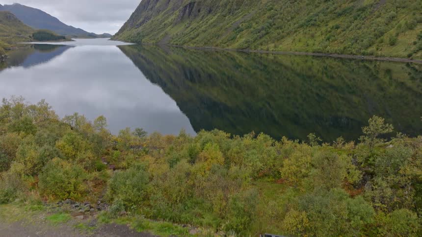 Aerial view of a gray overlanding camper van parked on a cliffside overlooking a serene mountain lake with perfect reflections