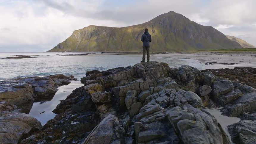 A lone hiker stands on a rocky outcrop on a scenic beach, admiring the vast mountain landscape and calm ocean