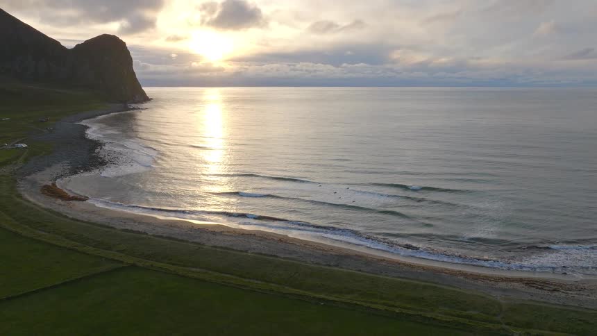 A tranquil aerial shot captures a golden sunset over a calm ocean, highlighting a beautiful secluded beach next to lush green coastal fields