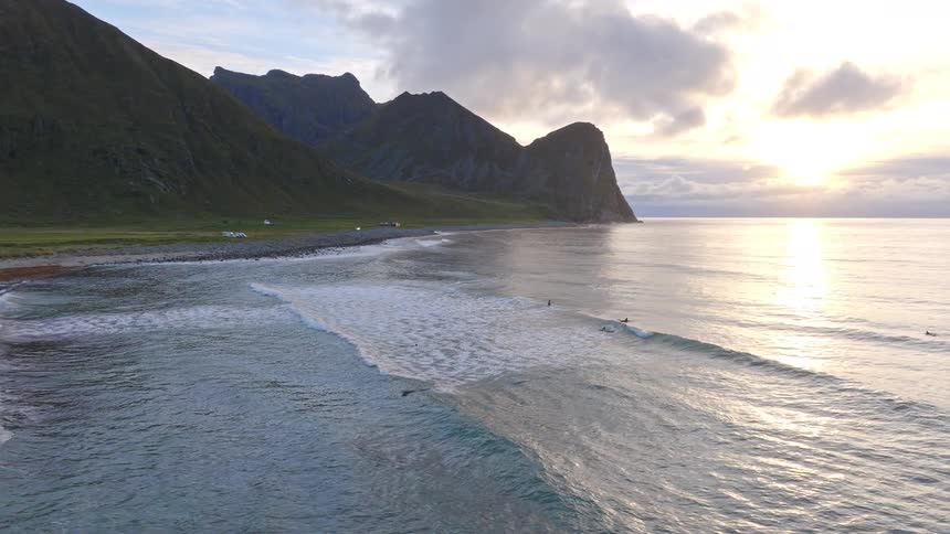 A stunning aerial shot of surfers enjoying the waves on a scenic coastline with majestic mountains during a beautiful sunset