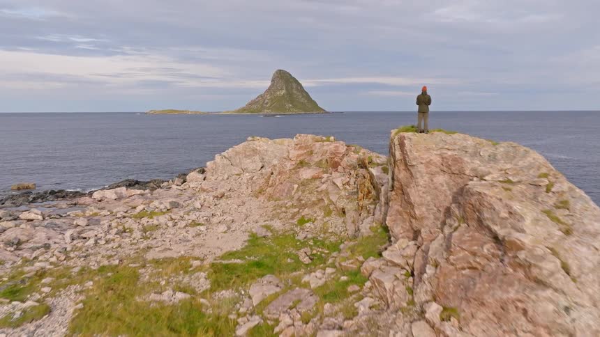 An aerial view slowly pushes in on a solitary figure standing atop a rugged coastal cliff, gazing out at the vast ocean and a dramatic island on the horizon
