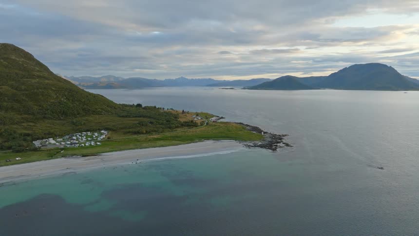 An expansive aerial shot captures a scenic coastline in Norway, featuring a pristine beach, clear turquoise water, and a campground against a backdrop of towering mountains
