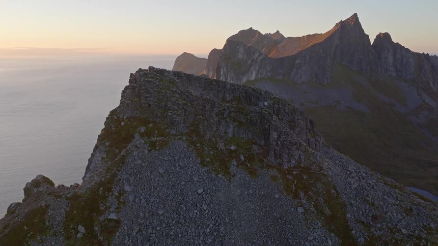 A slow aerial reveal of a majestic, rocky mountain ridge overlooking the vast ocean during a beautiful golden hour sunset