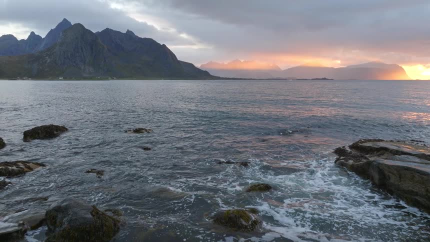 An aerial view slowly advances over a rocky sea coast as a dramatic sunset glows behind majestic mountains
