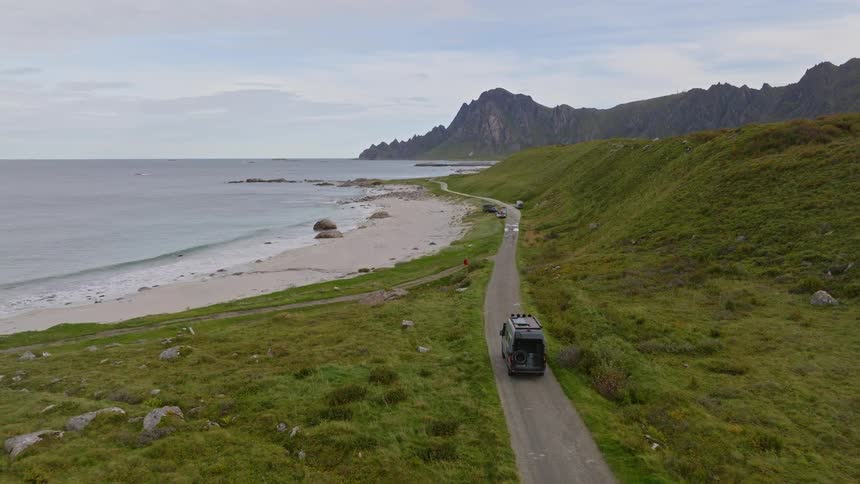 An aerial view follows a camper van driving along a scenic coastal road next to a sandy beach and green hills in Norway