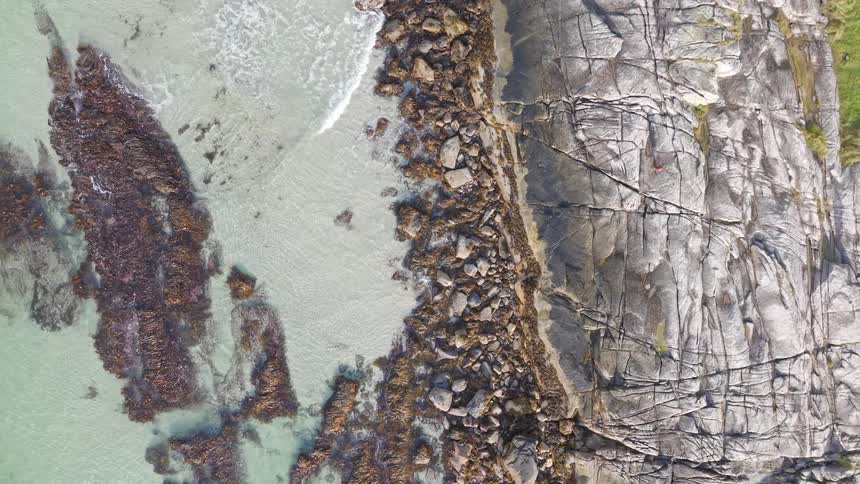 Aerial top-down view rises above a person relaxing on a rocky coast next to the clear turquoise ocean
