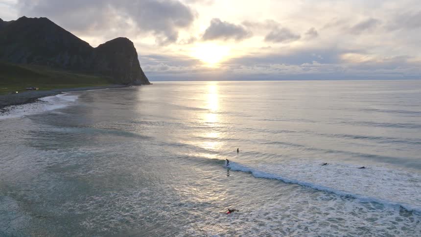 A serene slow-motion aerial follows surfers enjoying the waves against a stunning mountainous coastline during a golden sunset