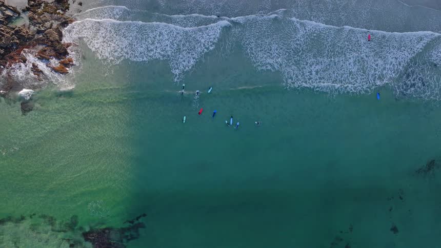 A serene top-down aerial view shows a group of surfers with colorful boards floating in the clear turquoise ocean as gentle waves break on the shore