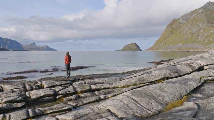 A solitary person stands on a rocky shoreline, peacefully observing the vast and serene mountain landscape over the water