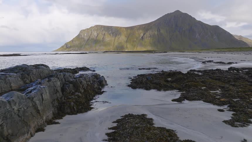 Aerial footage pulls back to reveal a lone man walking along a scenic, rugged beach with a towering mountain in the background
