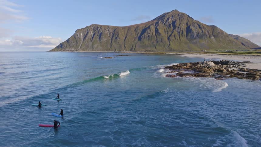 An aerial pullback reveals a group of surfers enjoying the tranquil waves at a stunning beach below a large mountain