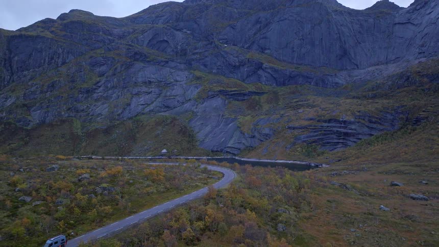 An aerial view captures a lone camper van driving on a winding road through a majestic and dramatic mountain valley on an overcast day