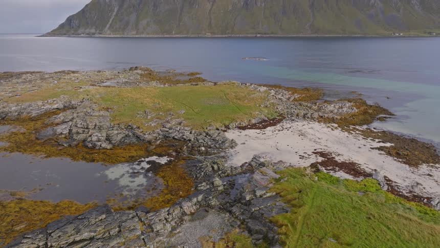 Aerial view tilts up from a rocky islet with a sandy patch to reveal a massive mountain across the tranquil fjord