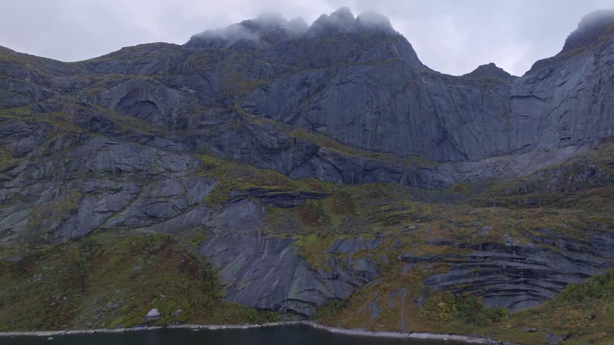 An aerial view reveals a vast, rugged mountain range rising from a dark fjord on a moody, overcast day