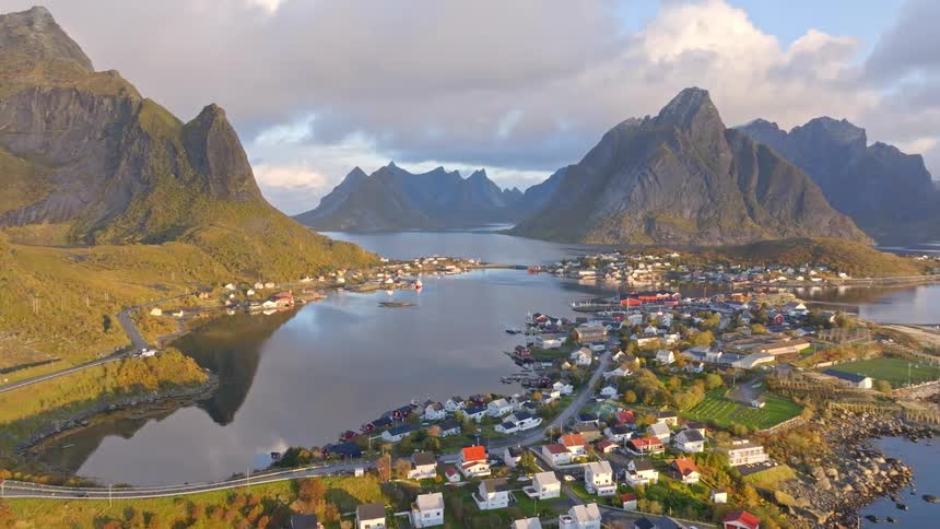An expansive aerial shot pans over a picturesque Norwegian coastal village nestled between majestic mountains and a calm, reflective fjord during golden hour