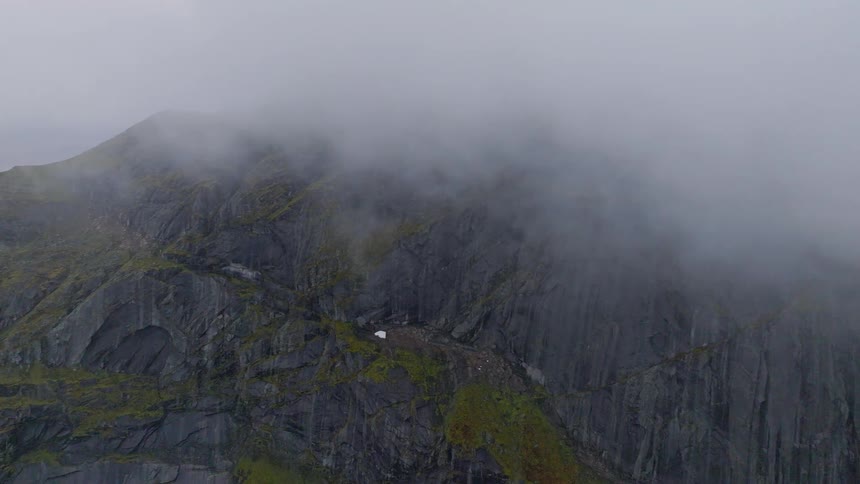 An epic aerial shot reveals a vast, rocky mountain as thick fog rolls across its mossy slopes