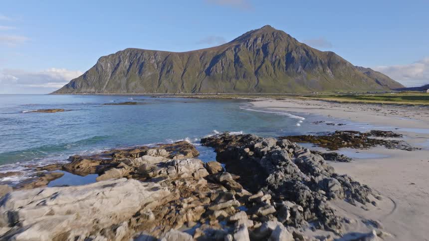 An aerial view glides over a stunning rocky beach toward the turquoise ocean with a majestic mountain backdrop in Lofoten, Norway
