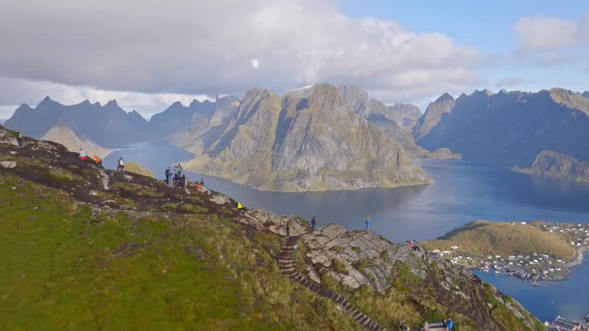 A stunning aerial pullback reveals hikers on a mountain summit enjoying the majestic view of a coastal village and fjord in the Lofoten Islands, Norway