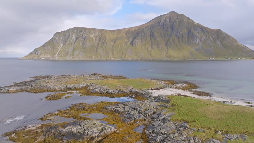 Aerial view reveals a drone pilot on a rugged, rocky island coast with a massive mountain in the distance