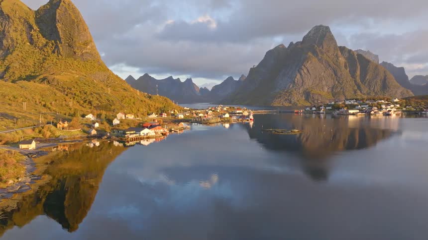 An idyllic aerial pullback reveals the stunning village of Reine in the Lofoten Islands, where majestic mountains reflect in the calm waters of the fjord at sunrise