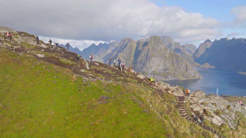 A beautiful drone shot follows hikers up a steep mountain trail, revealing a stunning panoramic vista of the fjords and town of Reine in Lofoten, Norway