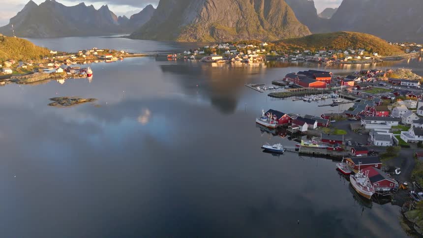 An aerial view pans up over the calm, reflective waters of a fjord to reveal the picturesque fishing village of Reine in the Lofoten Islands, Norway, at sunrise
