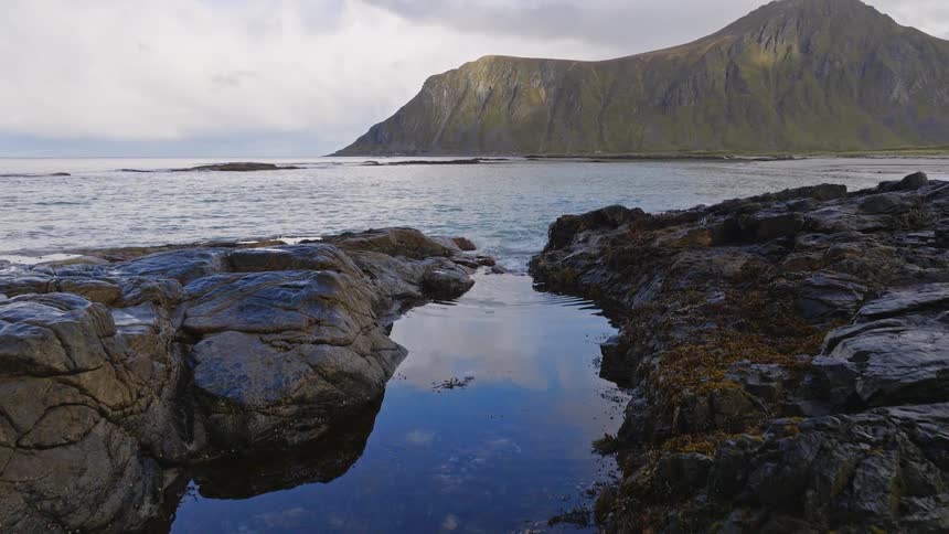 A tranquil tide pool on a rugged Scandinavian shoreline reflects the sky as the camera slowly reveals a majestic mountain across the bay