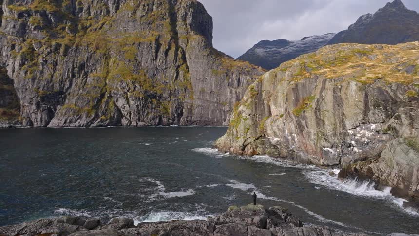 An epic slow zoom-out reveals a solitary person standing on a rocky shore, dwarfed by the immense cliffs and churning ocean of a Norwegian fjord
