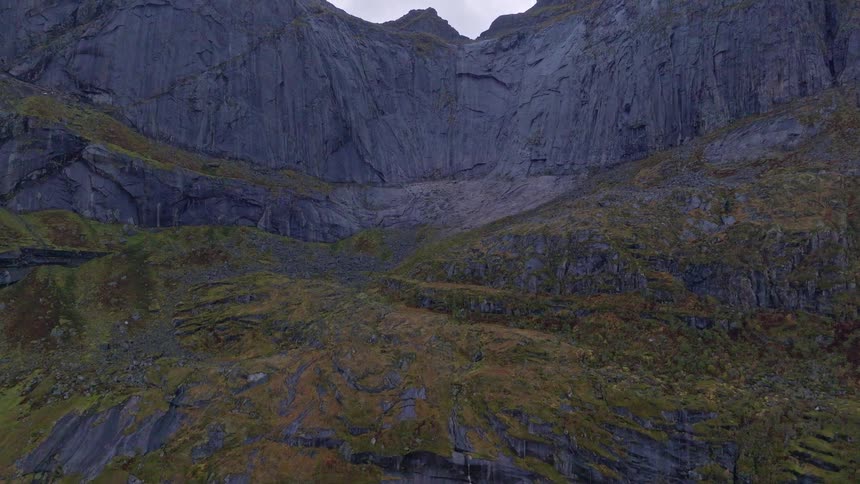 A dramatic aerial view tilts up from the shoreline to reveal a vast, rugged mountain face with autumn colors and cascading waterfalls