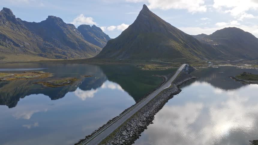 Ascending aerial shot of a stunning road and bridge crossing a tranquil lake, perfectly reflecting the majestic mountains of Lofoten, Norway