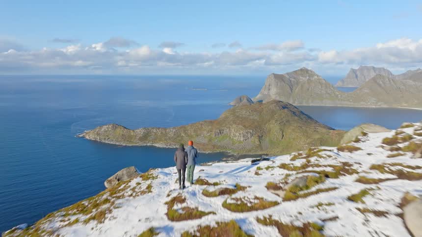 An adventurous couple stands together on a snowy mountaintop, taking in the spectacular aerial view of the ocean and coastal islands