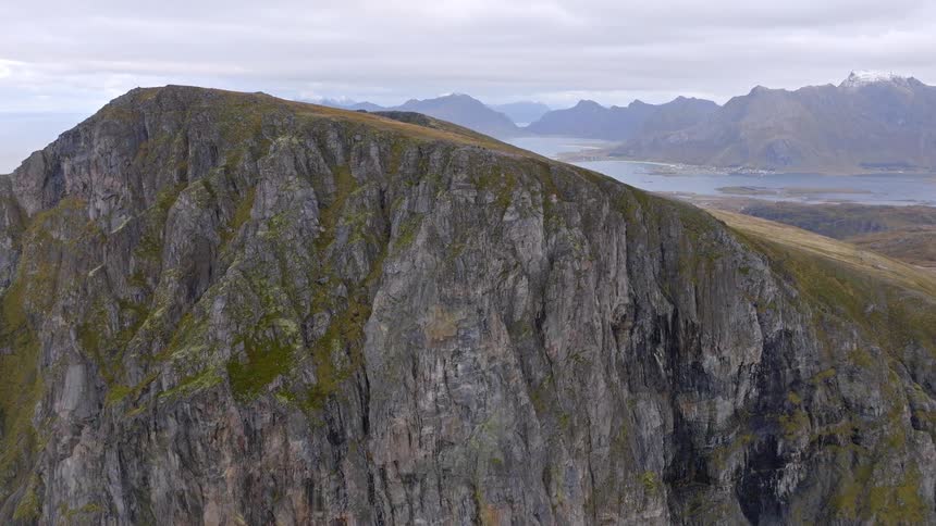 An aerial ascent reveals a vast, rugged mountain range overlooking a fjord and coastal villages in the distance