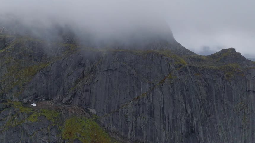 A dramatic static shot shows thick fog rolling over the top of a rugged, dark mountain face