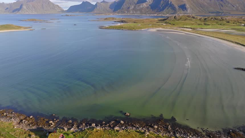 A breathtaking aerial flight over a serene turquoise bay and sandy beach, slowly revealing a majestic mountain landscape on the Norwegian coast