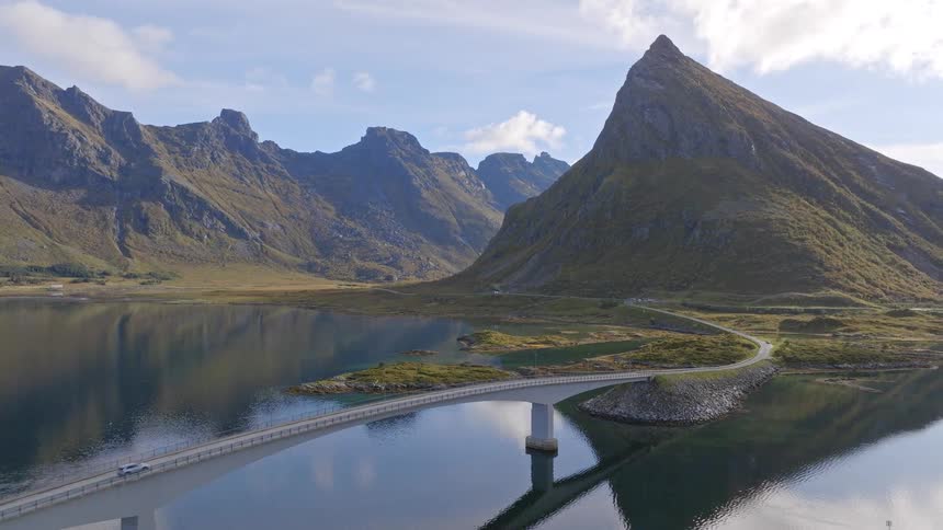 Scenic aerial pullback reveals a modern bridge crossing a tranquil fjord with majestic, reflecting mountains in Lofoten, Norway