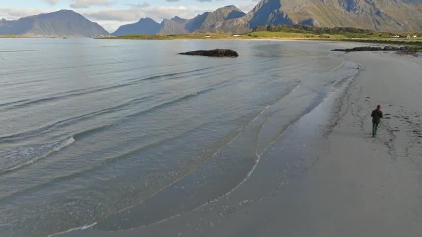 An aerial view follows a lone person walking along a sandy shoreline, tilting up to reveal a majestic mountain range across the ocean