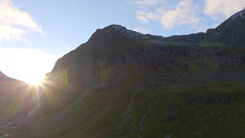 A majestic time-lapse captures the sun rising from behind a rugged, snow-dusted mountain, creating a brilliant sun flare